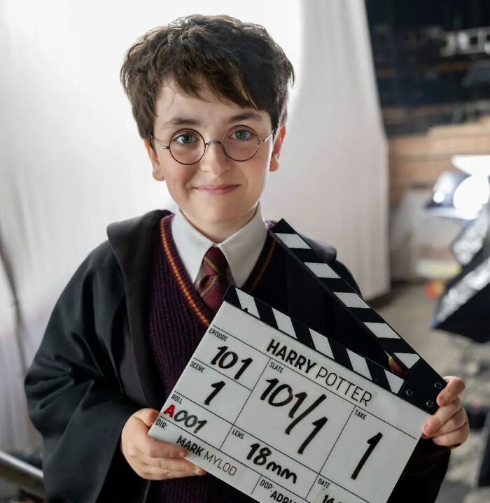 A young boy dressed as Harry Potter, wearing glasses and a Hogwarts uniform, holds a film slate for the Harry Potter series.