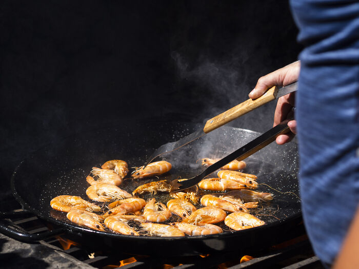 A person using tongs to flip cooking shrimp in a large pan over an open flame, with smoke rising. These shrimp might be part of unusual house rules for a family.