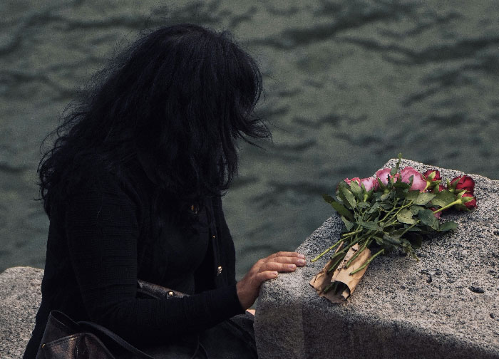 A woman with dark hair sits by water, next to a bouquet of roses, reflecting on the intriguing mysteries of WWII.
