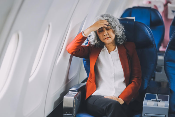A woman with gray hair in a red blazer clutches her head on an airplane during a family trip. A woman with gray hair in a red blazer clutches her head on an airplane during a family trip.