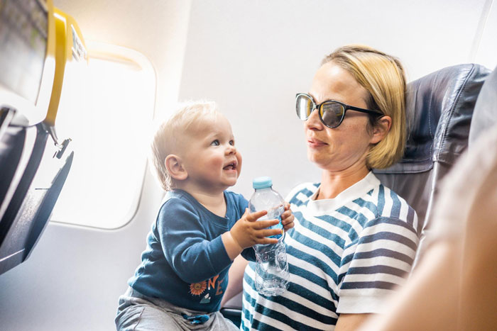 A mother wearing sunglasses holds a baby on her lap during a flight, with the baby holding a water bottle, impacting MIL and SIL. A mother wearing sunglasses holds a baby on her lap during a flight, with the baby holding a water bottle, impacting MIL and SIL.