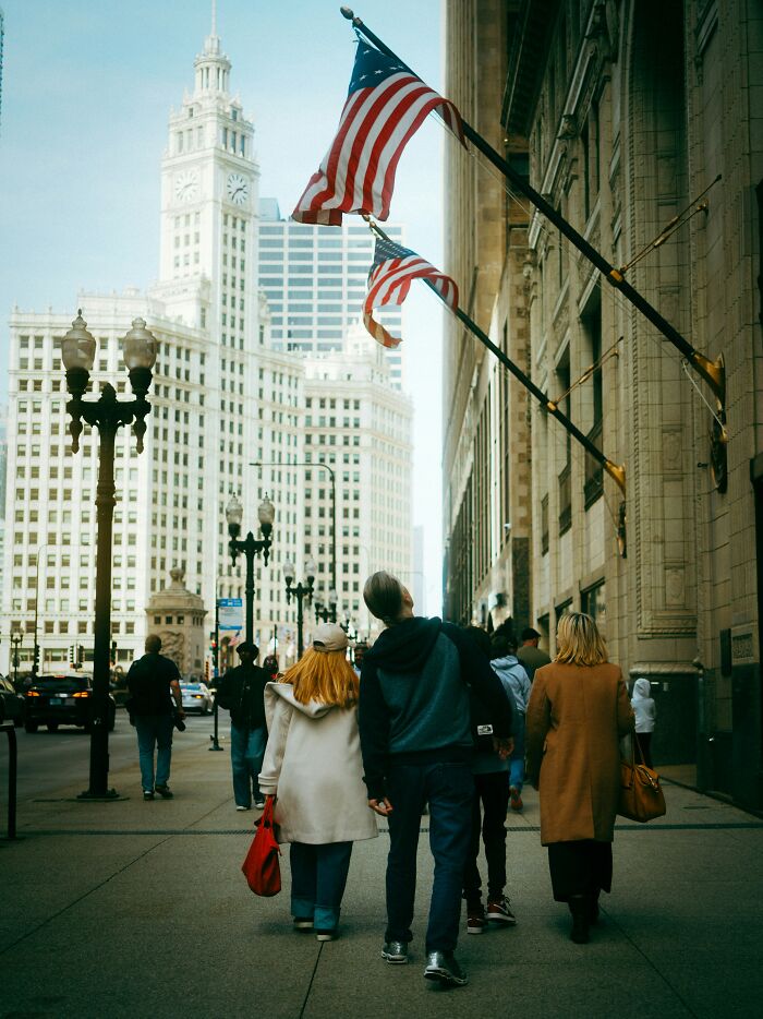 Newcomers in America walking past buildings with US flags, surprised by the sights.