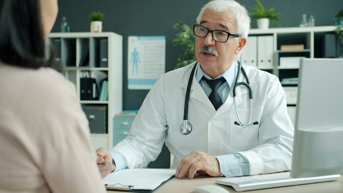 A male doctor in a white coat with a stethoscope talks to a patient in an office setting, reflecting medical consultation.
