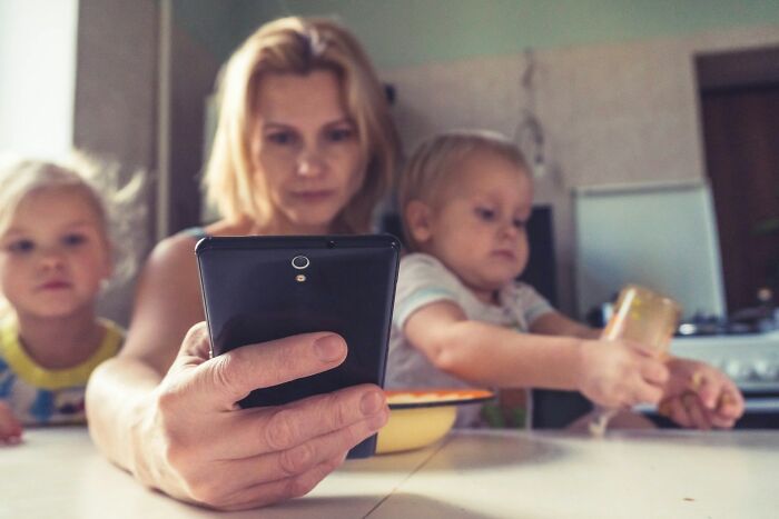 A parent distracted by their phone while two young children are nearby, one eating. This depicts a horrible parent scenario.