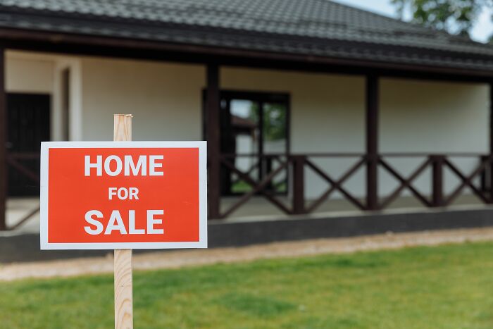 A 'HOME FOR SALE' sign in front of a house, representing the escape from cults and new beginnings.