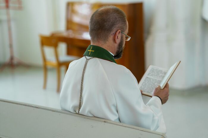 A man in clerical attire reading a book, viewed from behind, emphasizing the teachings and scriptures in cults.