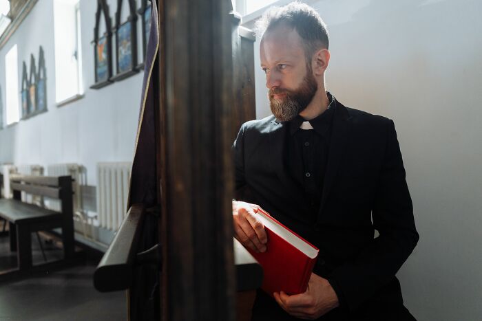 A man with a beard wearing a clerical collar and holding a red book, representing the religious figures in cults.