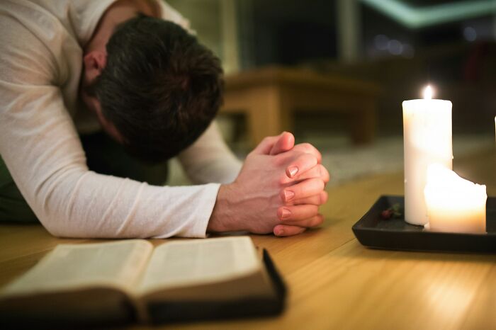 A man on his knees, head bowed, with clasped hands, praying near an open book and candles. This image relates to cults and ADHD.