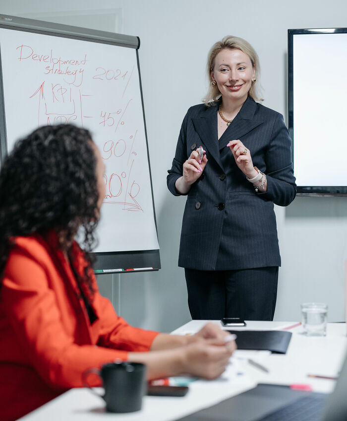 A woman giving a presentation in an office, smiling, while a coworker listens. The presentation avoids spicy personal details.