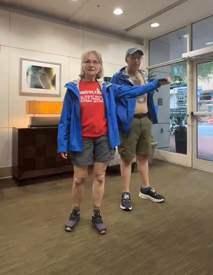 A white couple stands, a man in khaki shorts and a blue jacket and a woman in gray shorts, red shirt, and blue jacket, pointing in an apartment building. A white couple stands, a man in khaki shorts and a blue jacket and a woman in gray shorts, red shirt, and blue jacket, pointing in an apartment building.