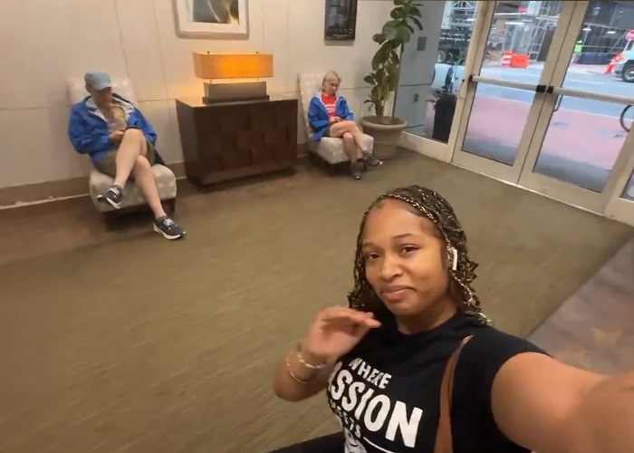 A Black hairstylist in an apartment building lobby, facing the camera with two white people in the background. A Black hairstylist in an apartment building lobby, facing the camera with two white people in the background.