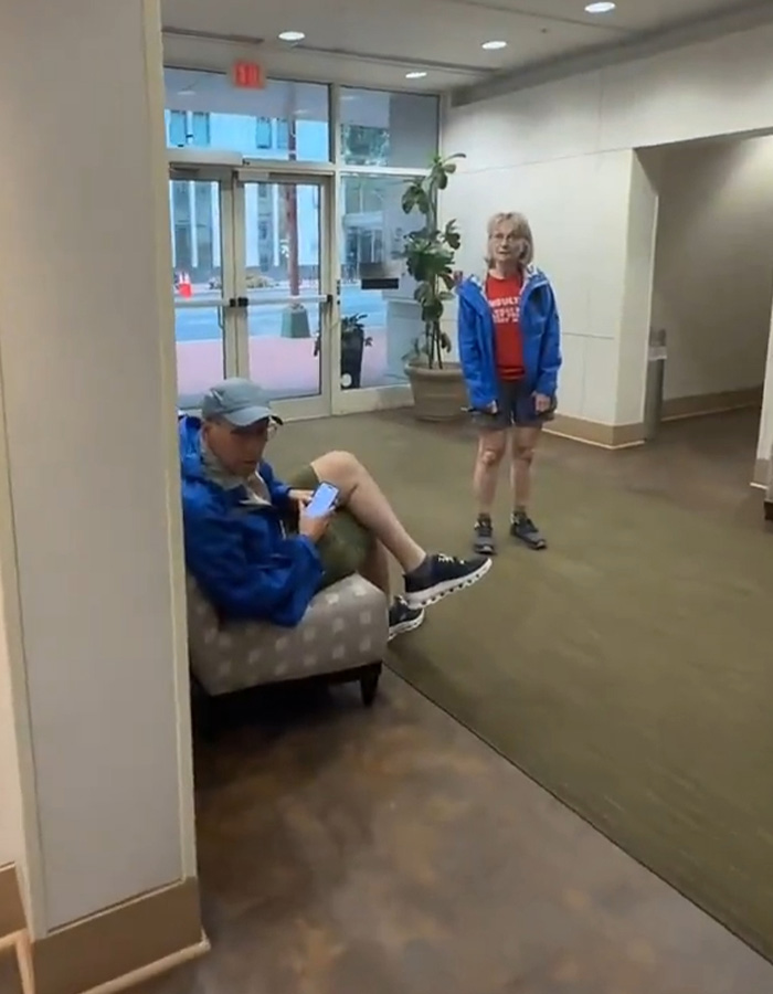 A white couple in an apartment building lobby; the man is seated using his phone, the woman stands nearby. A white couple in an apartment building lobby; the man is seated using his phone, the woman stands nearby.