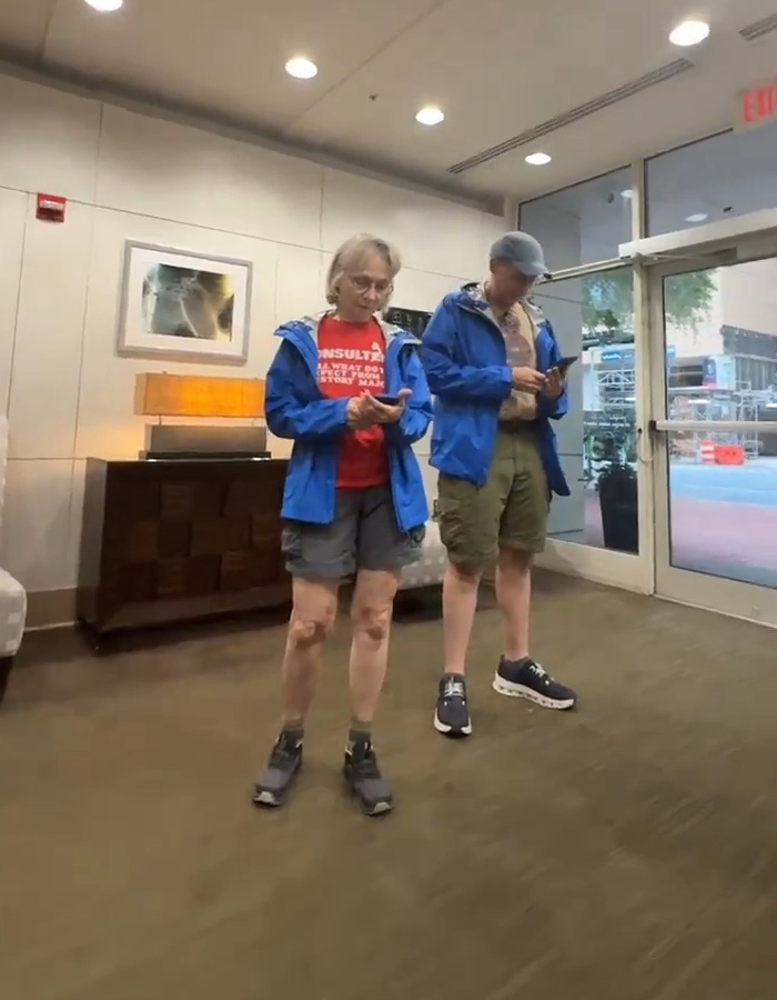 A white couple stands, a man in khaki shorts and a blue jacket and a woman in gray shorts, red shirt, and blue jacket, looking at their phones. An apartment building. A white couple stands, a man in khaki shorts and a blue jacket and a woman in gray shorts, red shirt, and blue jacket, looking at their phones. An apartment building.