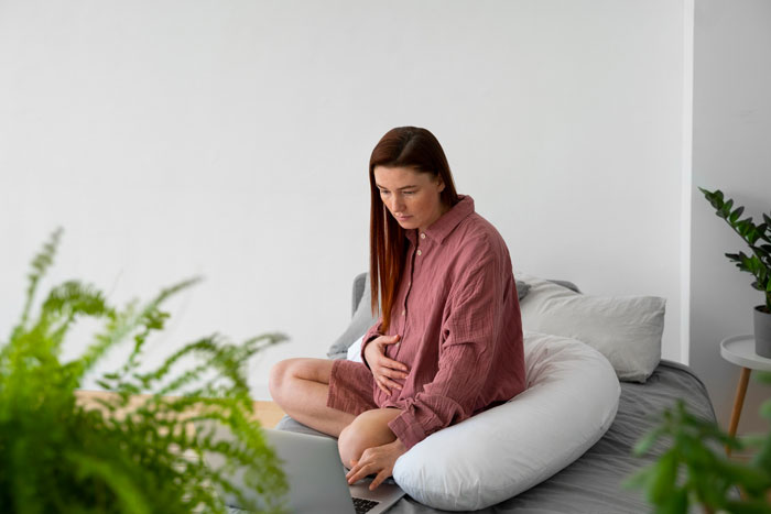 A pregnant mom sitting on a bed, looking at her laptop, contemplating decisions about kid's education and wardrobe rights.