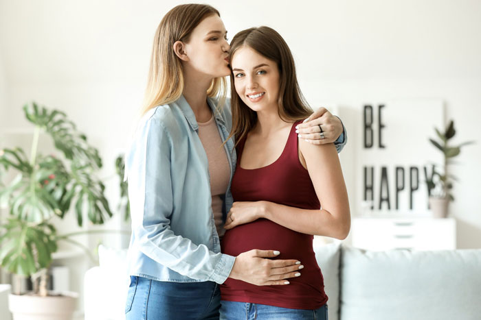 A pregnant woman smiles while her wife kisses her forehead, showing their love and a future kid's education.
