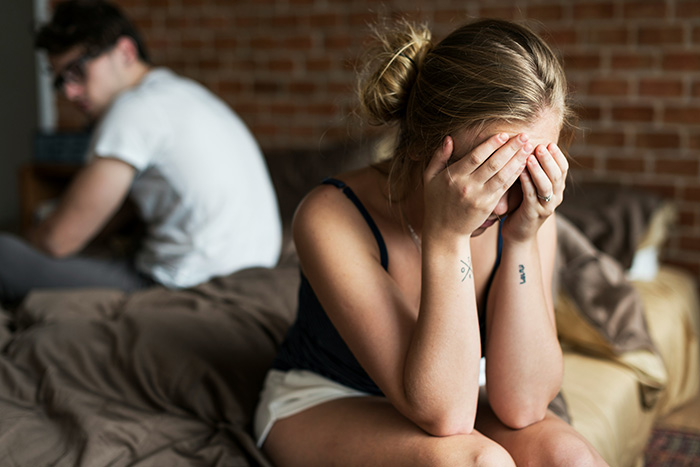 A woman crying with her face in her hands, as her husband sits on the bed, looking away after a horrible miscommunication.