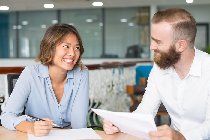 A woman with braces smiling and looking at a man holding papers. They are in a modern office setting. Epic comebacks.