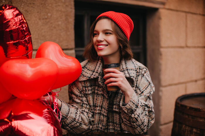 A woman holding heart balloons and a coffee cup, looking happy. She is wearing a plaid coat and a red beanie. Epic comebacks.