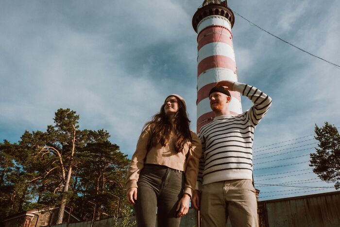 A couple stands in front of a striped lighthouse, looking up. Their epic comeback in life is their journey.