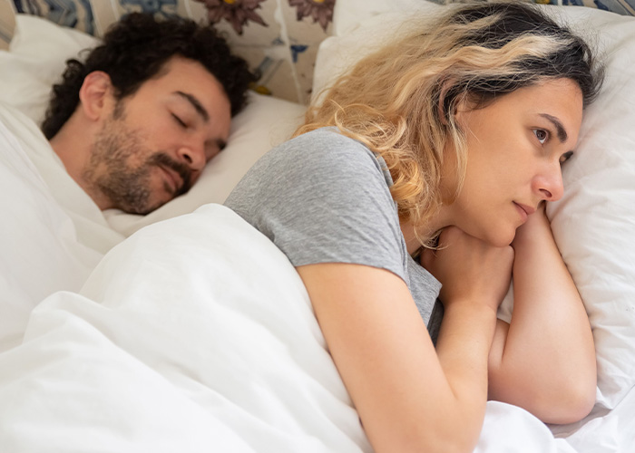 A woman looking sad in bed next to a sleeping man, depicting potential marital issues with separate apartments affecting marriage. A woman looking sad in bed next to a sleeping man, depicting potential marital issues with separate apartments affecting marriage.