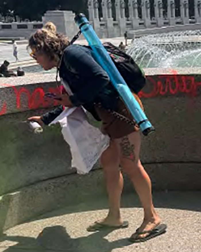 A woman vandalizing a WWII Memorial with red spray paint, carrying a blue tube and backpack.