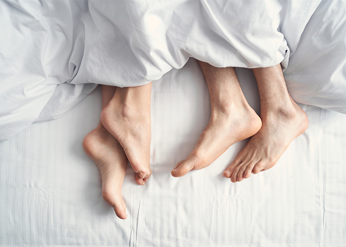 A couple's feet are visible under white sheets, illustrating the common question men ask in bed. A couple's feet are visible under white sheets, illustrating the common question men ask in bed.
