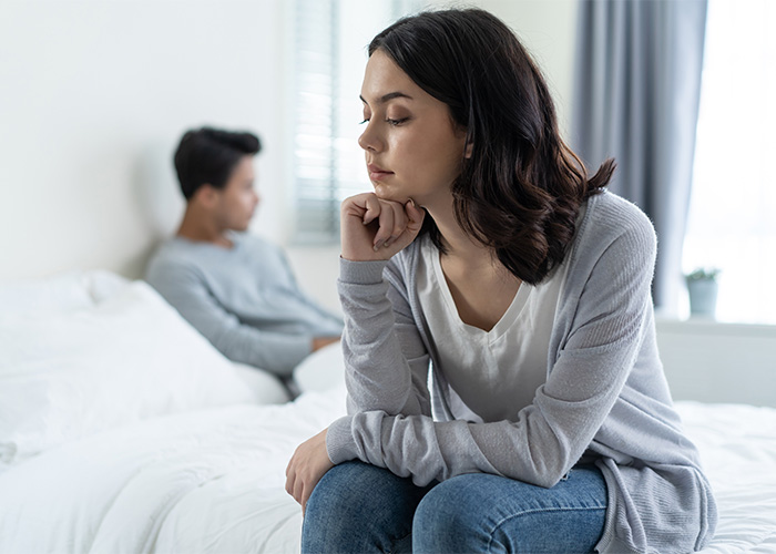 A woman sits on a bed looking upset, with a man blurred in the background, depicting mood killer questions in bed. A woman sits on a bed looking upset, with a man blurred in the background, depicting mood killer questions in bed.
