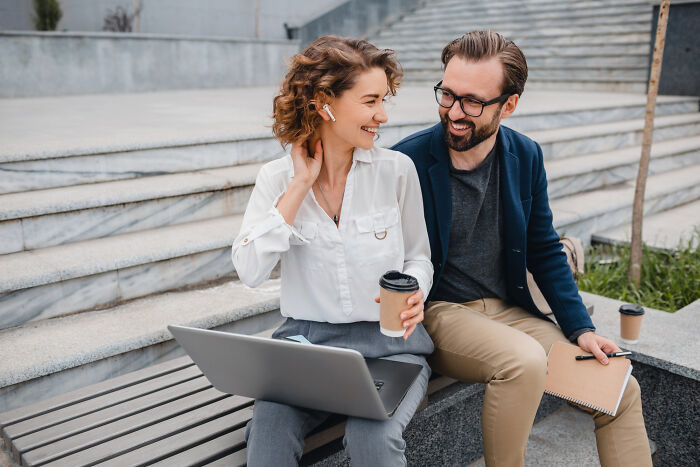 A man and a woman laughing while looking at a laptop outdoors, potentially discussing date nights or an escape.