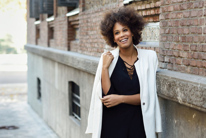 A smiling woman with curly hair leaning against a brick wall, feeling confident despite comments from women.