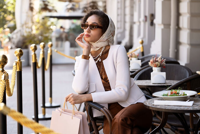 A stylish woman with sunglasses and a headscarf, holding shopping bags at an outdoor cafe, representing women's comments.