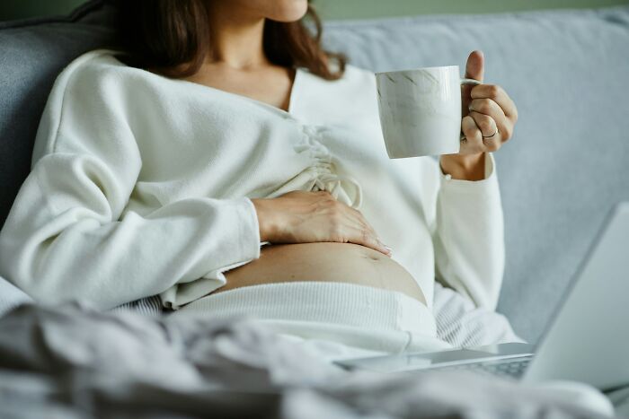 Pregnant woman on a couch with a laptop, holding a mug, contemplating worst things she endured.