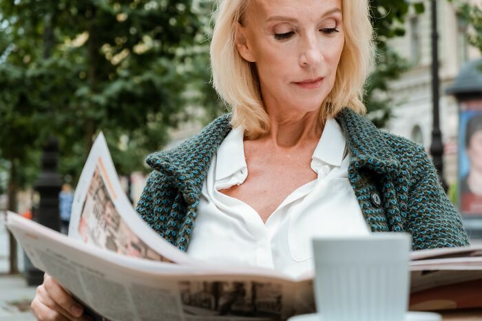 An older woman reading a newspaper at an outdoor cafe, pondering the worst things women put up with.