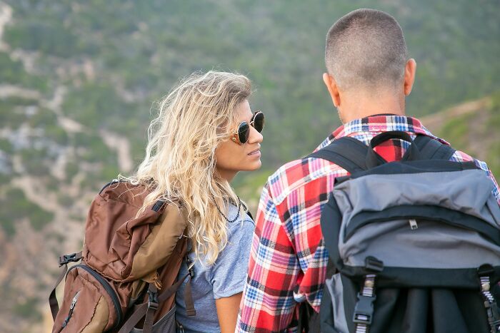 A woman wearing sunglasses and a backpack talks to a man, hiking. Women share what they put up with before walking away.