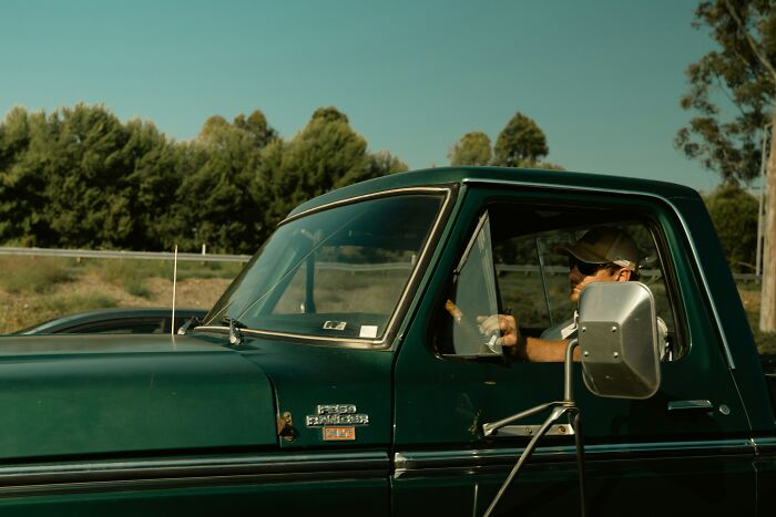 A man driving a vintage green Ford F-150 Ranger pickup truck against a backdrop of trees, illustrating things women put up with.