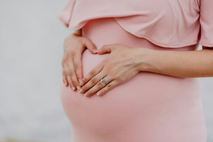 A pregnant woman in a pink dress forms a heart shape with her hands over her baby bump, illustrating a young and dumb decision.