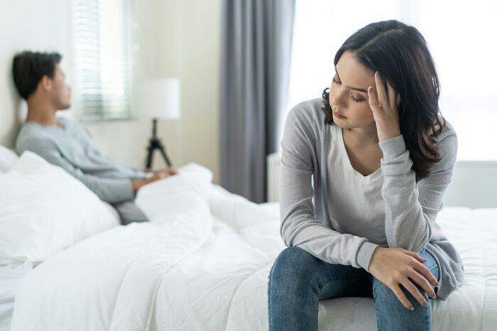 A woman sits on a bed, looking upset, with a man in the background, thinking about a young and dumb decision.