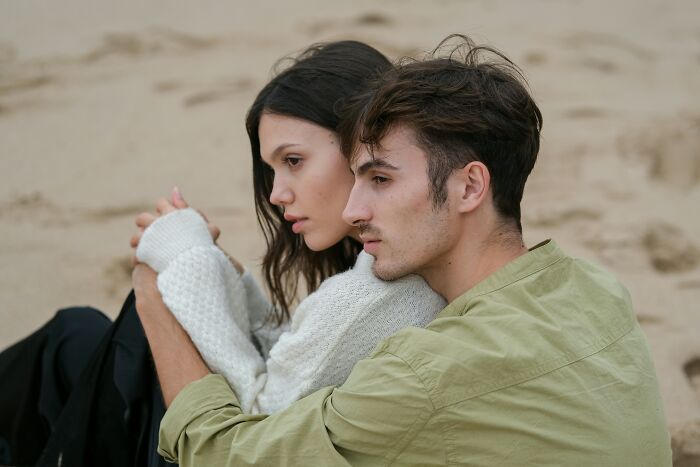 A couple embracing on the beach, with pensive expressions, recalling a young and dumb decision.