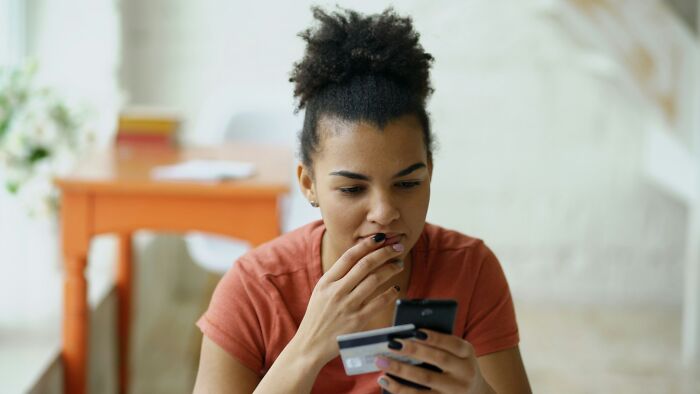 A woman looks at her phone with a credit card, contemplating a young and dumb decision.