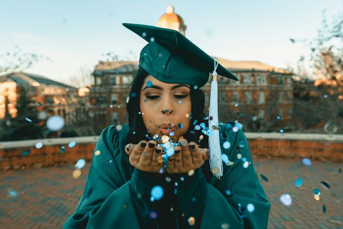 A graduate in a cap and gown blowing confetti from her hands, celebrating a young and dumb decision.