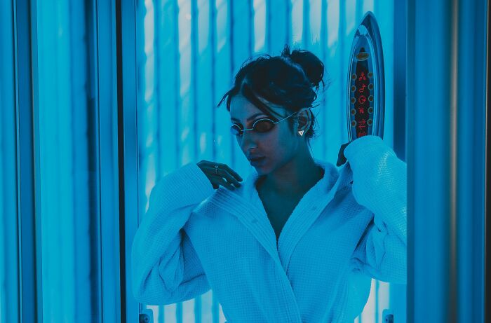 A woman in a white robe and sunglasses stands in a tanning booth, reflecting on a young and dumb decision.