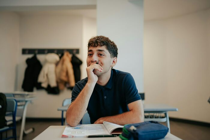A young male college student with curly hair and a nose ring sits at a desk, looking thoughtful during college life.