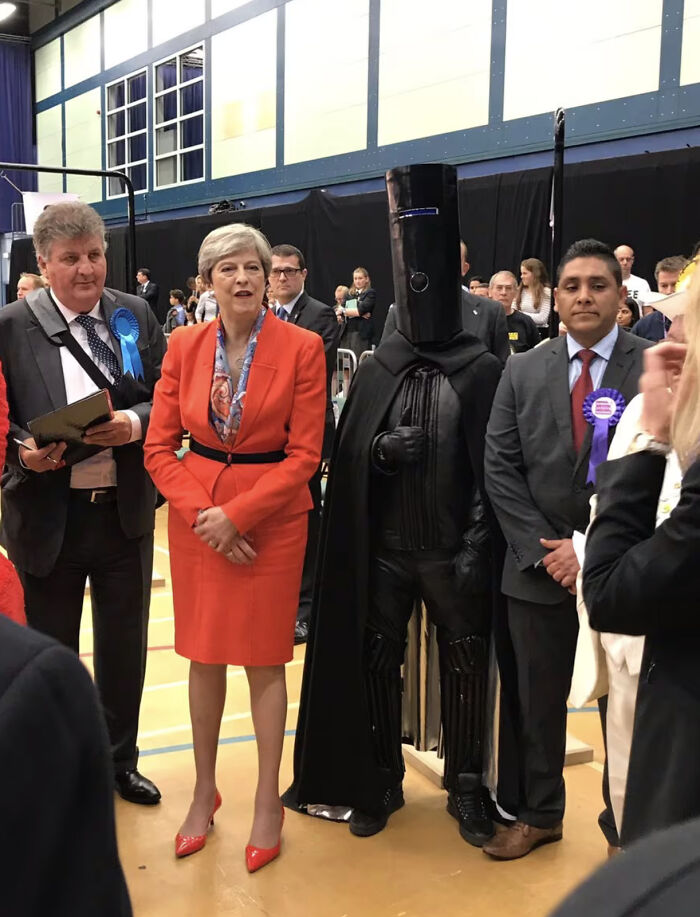 Theresa May next to a man in a black helmet costume at a polling station, an instance of unexpected British humor.