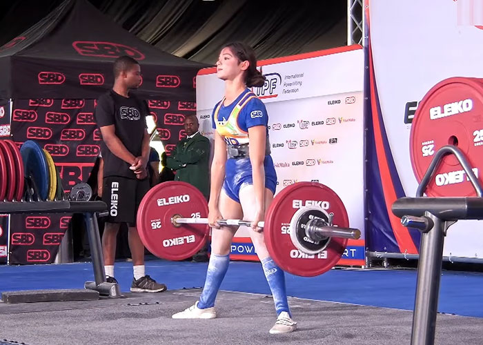 A female weightlifting medalist in a blue uniform, wearing a lifting belt, deadlifts a heavy barbell on a competition stage. A female weightlifting medalist in a blue uniform, wearing a lifting belt, deadlifts a heavy barbell on a competition stage.