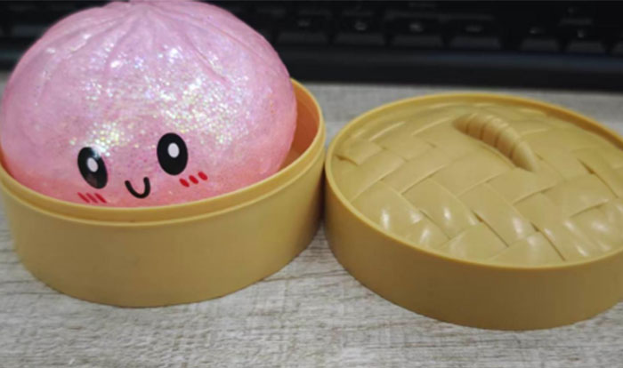 A pink glittery squishy toy with a smiling face inside a beige container, with the lid next to it, highlighting squishy toys. A pink glittery squishy toy with a smiling face inside a beige container, with the lid next to it, highlighting squishy toys.