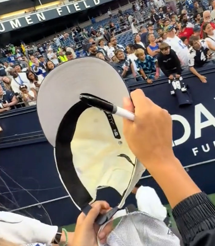 A hand holding a marker to sign a white NFL hat at a crowded stadium match A hand holding a marker to sign a white NFL hat at a crowded stadium match