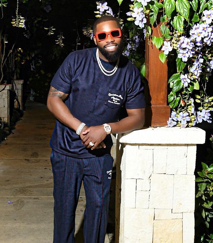Influencer in a dark blue outfit and red sunglasses, posing outdoors near flowers after a male cosmetic procedure.
