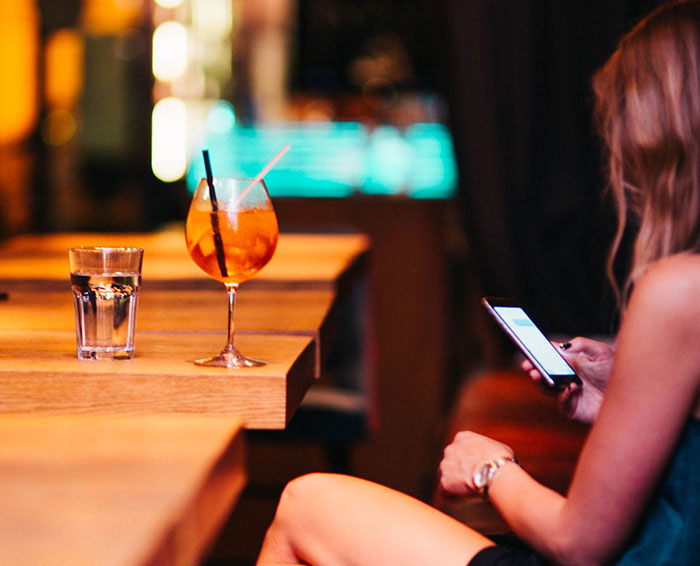 Woman in a bar holding a phone with a drink on the table, highlighting dating mistakes. Woman in a bar holding a phone with a drink on the table, highlighting dating mistakes.