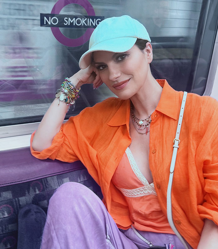 Laura Pausini in an orange shirt and cap, posing inside a train for an Instagram versus reality comparison.