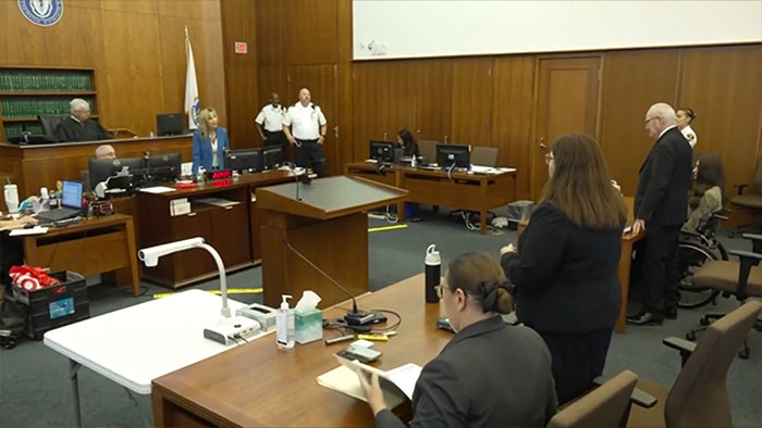 A wide shot of the courtroom reveals Lindsay Clancy in a wheelchair, surrounded by legal personnel and a standing jury. A wide shot of the courtroom reveals Lindsay Clancy in a wheelchair, surrounded by legal personnel and a standing jury.