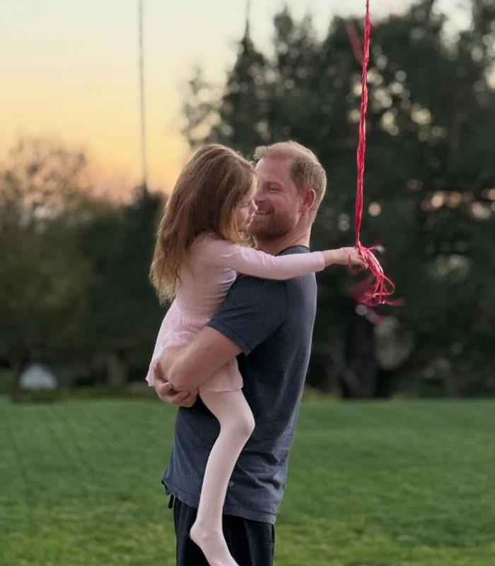 Prince Harry holding his daughter, who wears a pink dress and holds a red balloon, regarding kids' schooling.